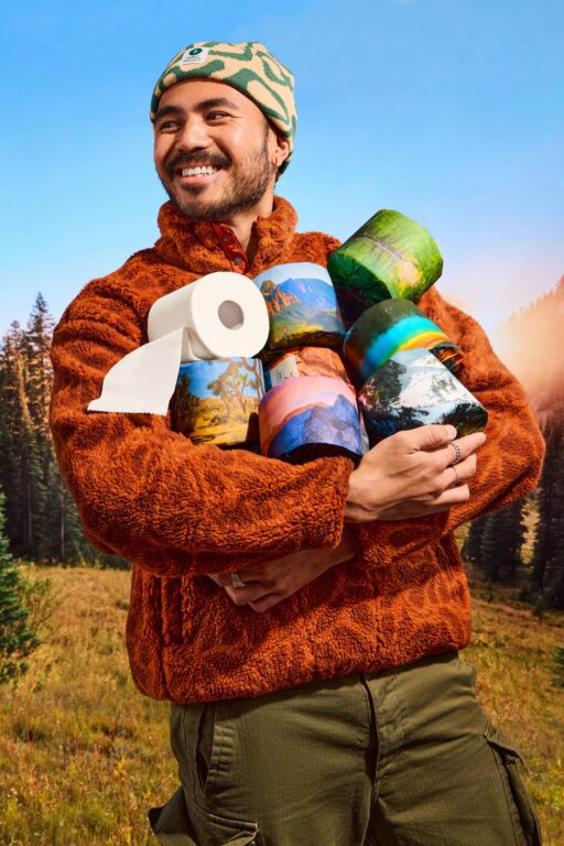 Man in orange fleece holding toilet paper rolls with scenic landscape designs.